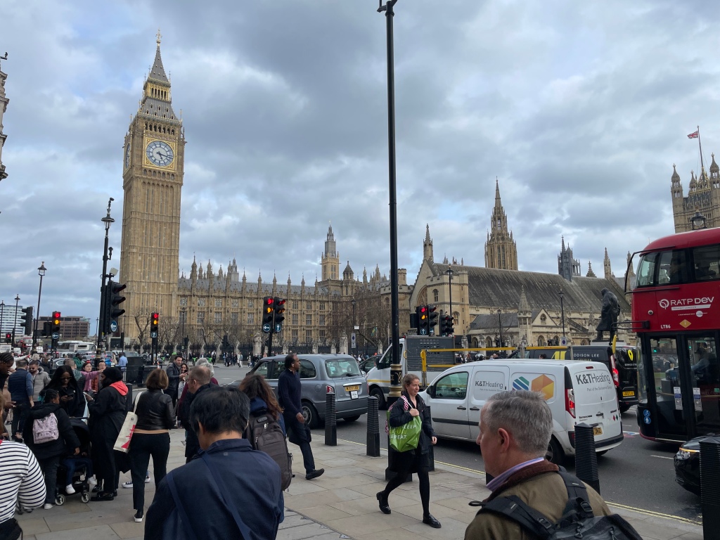 A picture of Parliament Square. In the background there are the Houses of Parliament and the Elizabeth Clock Tower (Big Ben). In the foreground are people walking on the pavement and lots of traffic